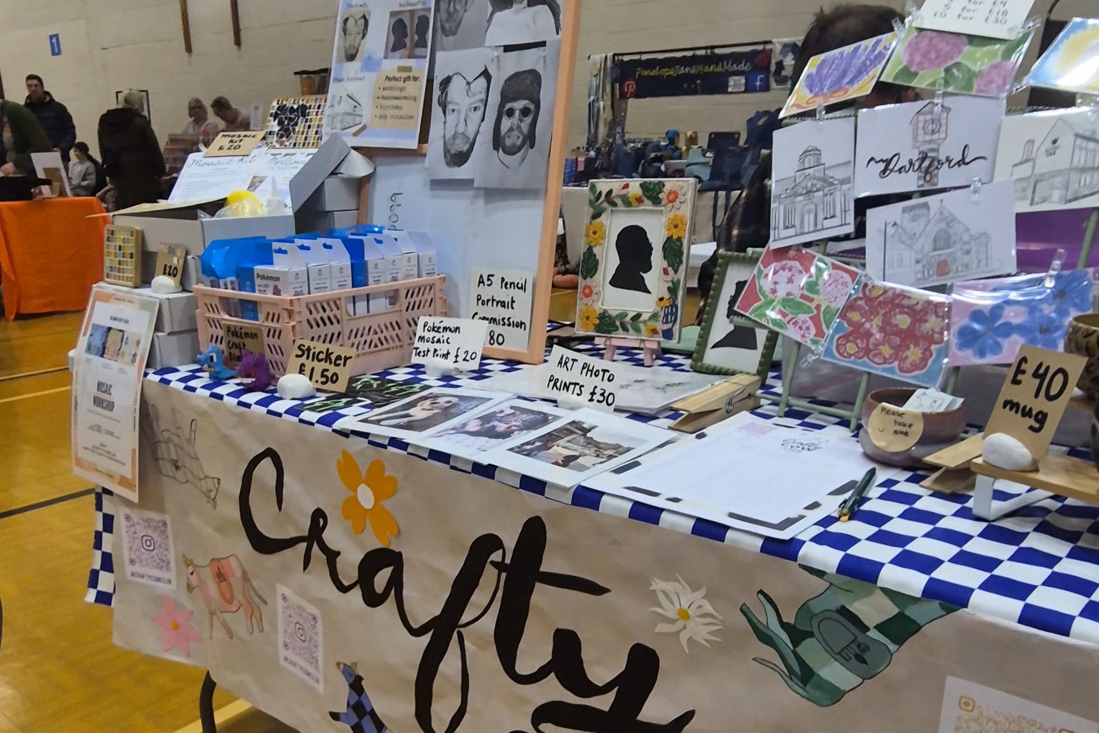 A craft booth table at an indoor market or fair, covered with handmade items, cards, and a large banner reading 'Crafty Cow'. The table is set in a gymnasium with wood floors and walls visible in the background.