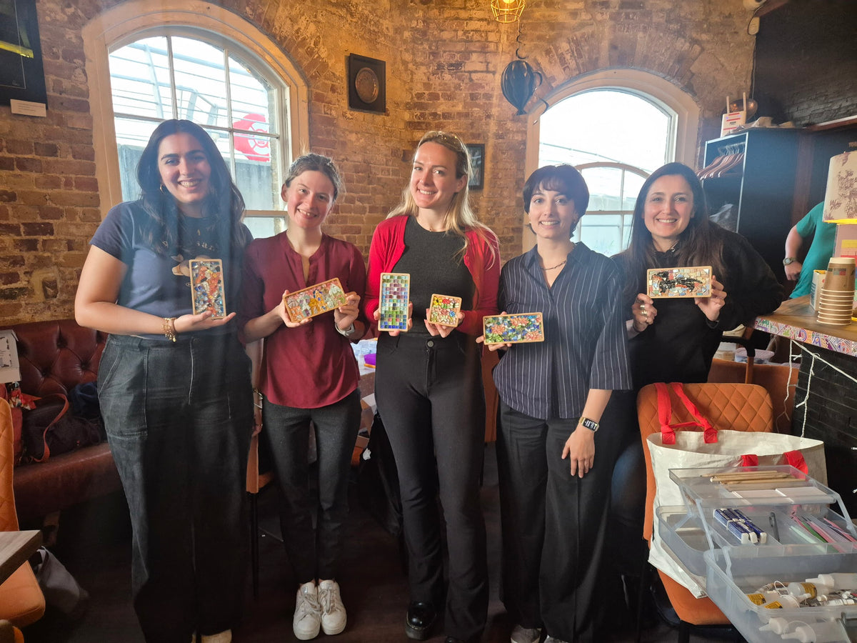 Five women standing together in a rustic indoor workshop or studio, each holding a small colorful rectangular craft or art piece. The room has exposed brick walls, large arched windows, and a table with supplies in the foreground.
