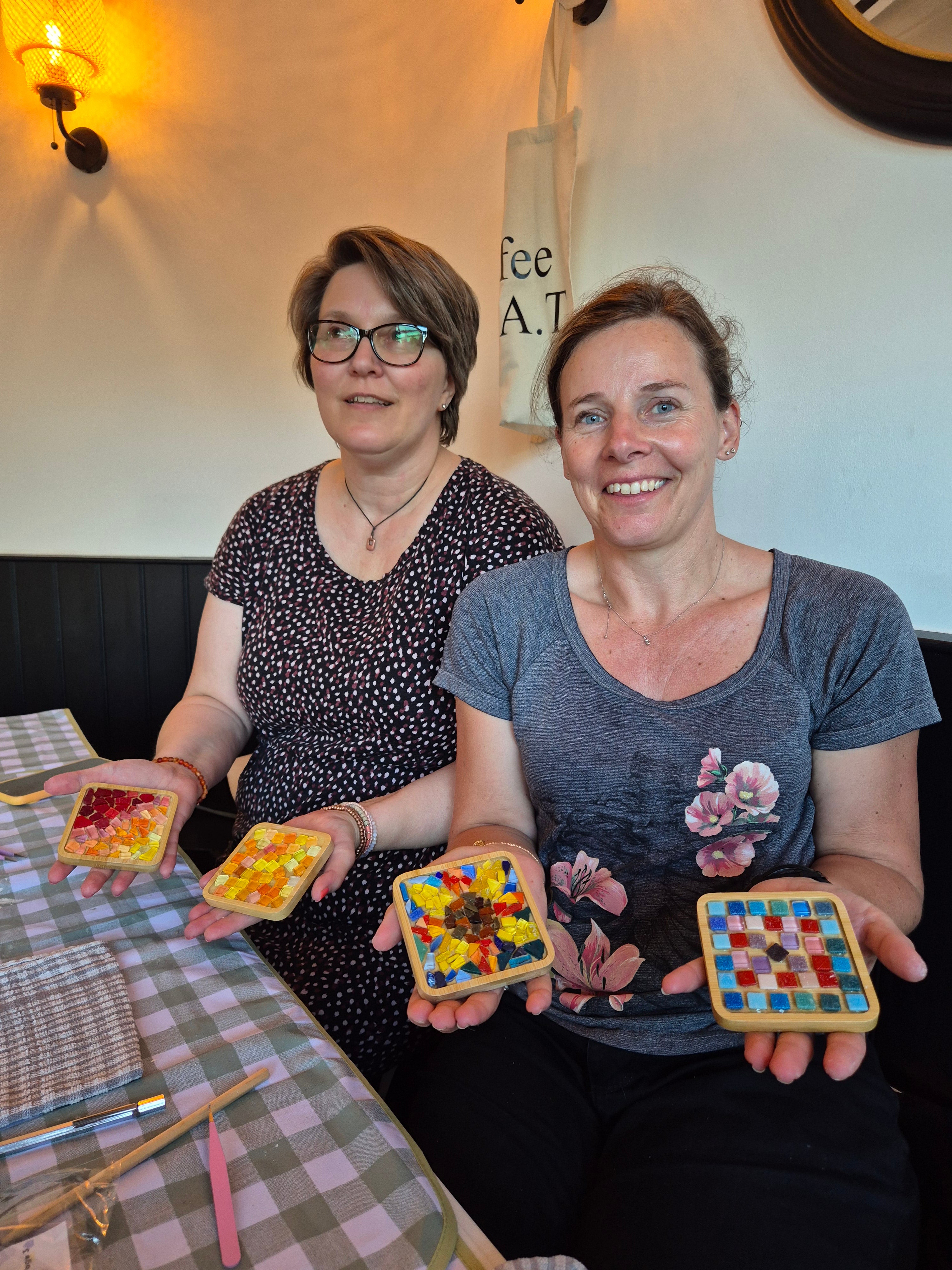 Two women holding colorful square objects with patterns, possibly mosaics or stained glass, in a casual indoor setting.