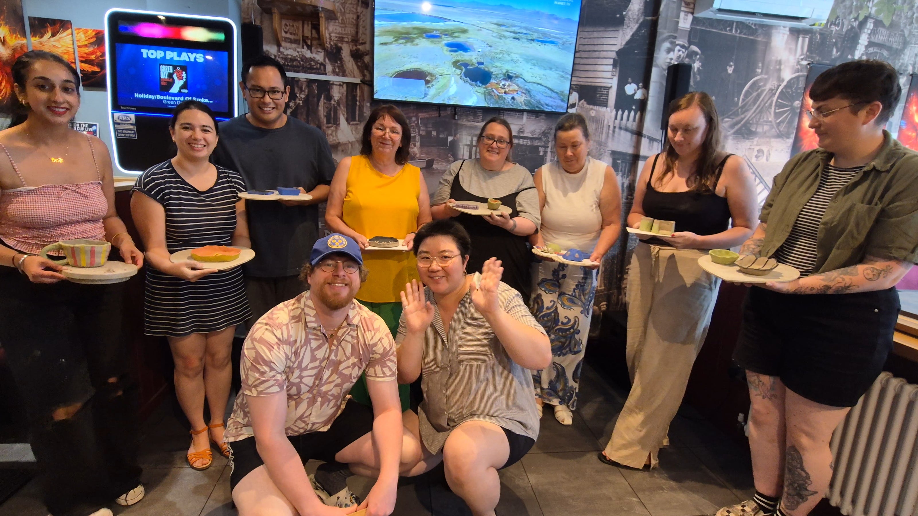 A group of ten people posing together in what appears to be a themed restaurant or event space, holding small plates with food. Several people are smiling and waving at the camera, with screens and posters visible in the background.