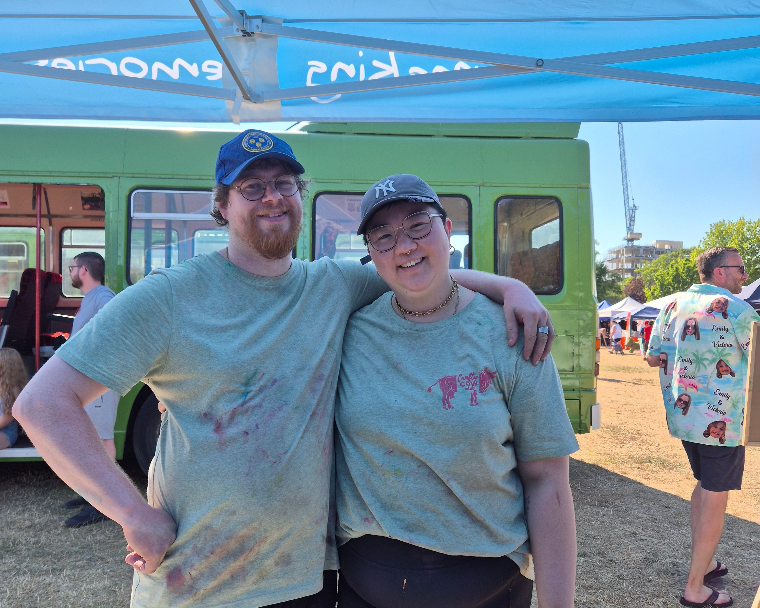 Two people standing outdoors under a light blue canopy tent, wearing light-colored t-shirts with pink and purple paint splatters, black shorts, and blue caps. A green vehicle is visible in the background. 