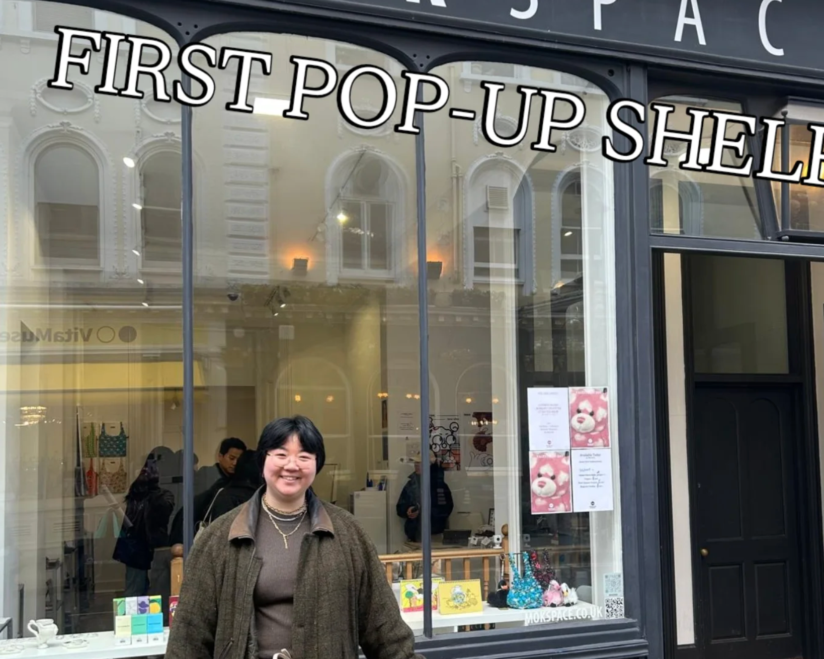 A woman stands smiling in front of a storefront with large windows and the sign 'MOKSPACE' and 'FIRST POP-UP SHELL' visible. The shop exterior is dark gray/black with white lettering, and the interior is warmly lit.